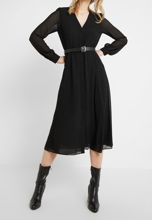 Woman wearing a long-sleeve black button-up midi dress with a black belt and black ankle boots, standing with hands on hips.