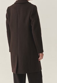 Langer brauner Wollmantel mit Kragen, gerader Schnitt und Rückenschlitz. Weiche Textur, minimalistisches Design.