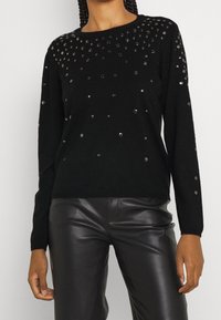 Pull noir en matière douce, orné de sequins ronds dispersés sur le devant, avec des manches longues et un col rond.