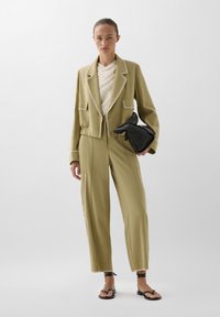 Costum verde măsliniu, ajustat, cu cusături albe contrastante, având un sacou scurt cu buzunare aplicate și pantaloni largi; poșetă neagră tip clutch.