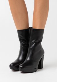 Botas de tobillo de cuero negro con punta redonda, textura suave, tacón alto cuadrado y panel lateral elástico para facilitar su uso.