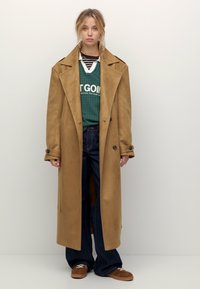 Jeune femme se tenant debout portant un long trench-coat beige, un pull vert superposé sur une chemise rayée, un jean foncé et des baskets marron.
