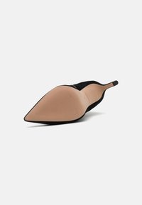 Zapato de tacón alto de ante negro con punta afilada y una suela beige lisa, que presenta surcos texturizados para mayor agarre.