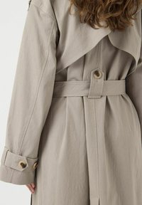 Vista trasera de un abrigo trench beige, con un cinturón en la cintura y una pestaña con botón, además de una correa de manga con botones, llevado por una persona con cabello largo y castaño.