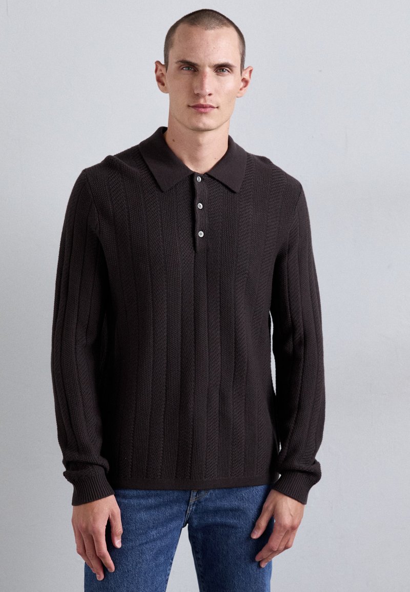 Temno rjav pleten pulover z rebrastim teksturami, dolgimi rokavi in zloženim ovratnikom. Ima tri gumbe na sprednjem pasu.