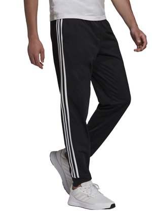 Pantalones deportivos negros con tres rayas blancas a cada lado, hechos de una tela suave, combinados con zapatillas deportivas blancas.
