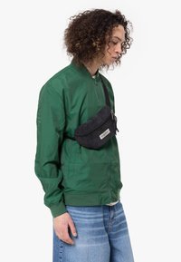 Giovane con capelli ricci indossa una giacca bomber verde, jeans azzurri chiari e una borsa a tracolla nera sul petto, mentre guarda verso il basso a destra.