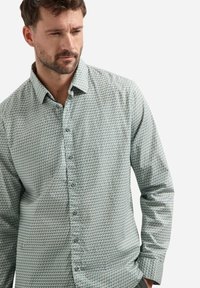 Man met kort bruin haar en een baard, gekleed in een overhemd met lange mouwen en een klein geometrisch patroon op een lichte achtergrond.