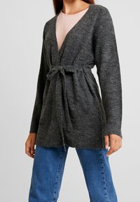 Kvinna iklädd en mörkgrå stickad cardigan knuten i midjan över en ljusrosa topp och blå jeans, stående mot en enkel bakgrund.