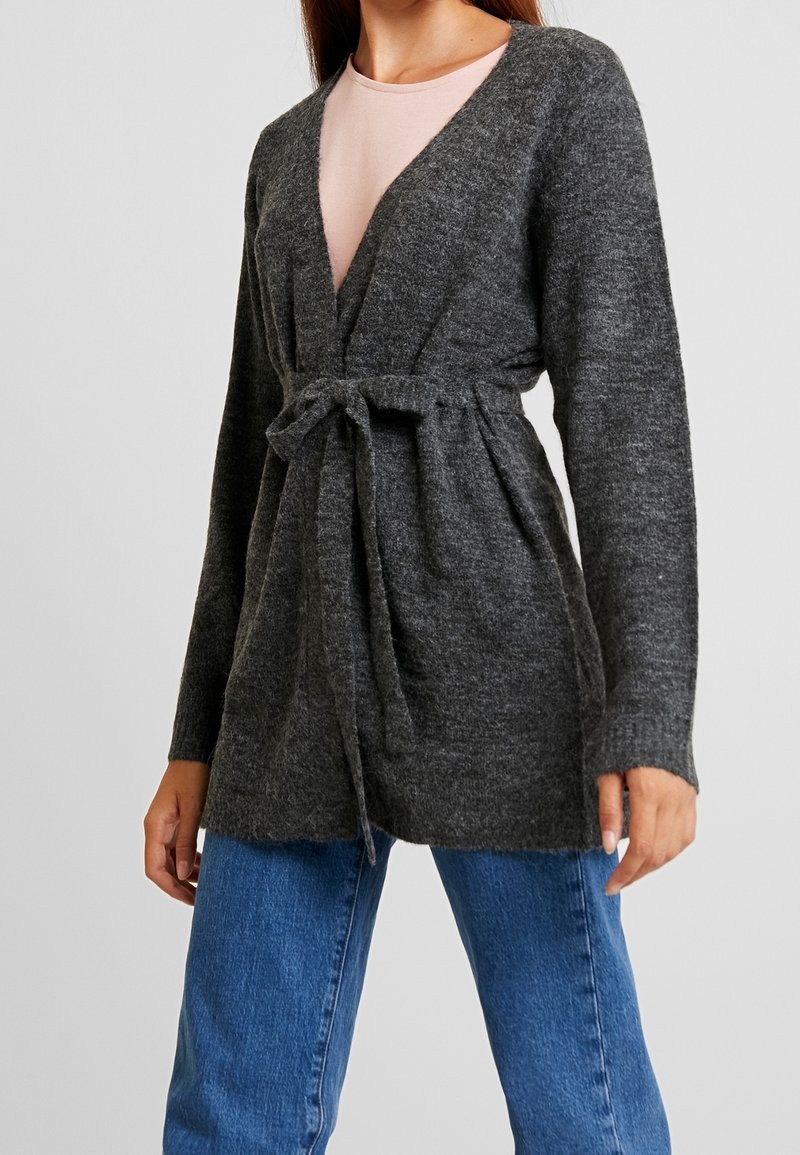 Kvinna iklädd en mörkgrå stickad cardigan knuten i midjan över en ljusrosa topp och blå jeans, stående mot en enkel bakgrund.