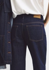 Jeans in denim blu scuro con cuciture arancioni, due tasche posteriori e un'etichetta logata color sabbia in vita. Accompagnati da una camicia in denim.