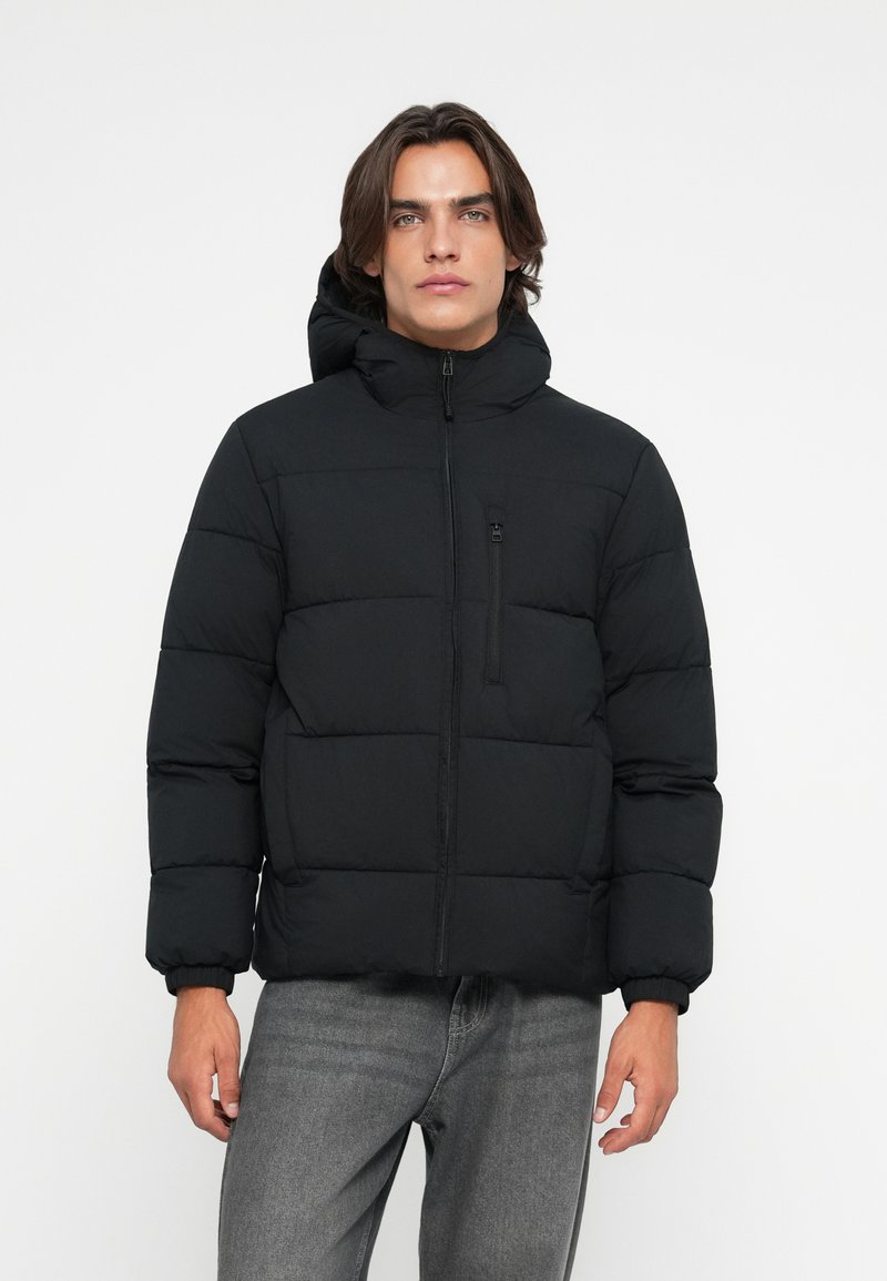 Uomo che indossa un giubbotto invernale nero con cappuccio e jeans grigi, in piedi davanti a uno sfondo bianco semplice.