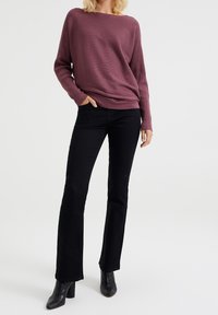 Nainen seisoo vasen käsi taskussa, päällä ribattu purppuranvärinen villapaita, mustat bootcut-farkut ja mustat nilkkurit valkoista taustaa vasten.