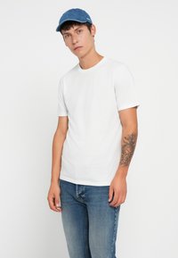 Biała koszulka z krótkim rękawem, materiał bawełniany, okrągły dekolt. Niebieska czapka z denimu, zakrzywiony daszek, z detalem logo. Model w jasnoniebieskich dżinsach.