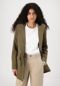 Giacca verde oliva con cappuccio, dotata di zip e bottoni a pressione, vita regolabile con cordino e molteplici tasche, indossata sopra una camicia bianca.