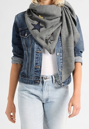 Femme portant un jean en denim clair, une veste en denim bleu et une grande écharpe grise ornée de patches en forme d'étoiles.