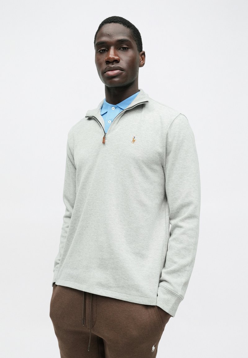 Helles graues Zip-up Sweatshirt mit einem blauen Hemd darunter. Verfügt über ein kleines Logo auf der linken Brust und hat einen lockeren Schnitt.