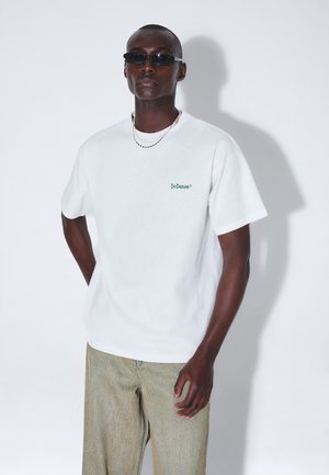 Mężczyzna noszący czarne okulary przeciwsłoneczne, białą koszulkę Dr.Denim, beżowe spodnie i srebrny naszyjnik z łańcuszka, stoi z jedną ręką za plecami.