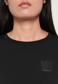Musta puuvillapaita, jossa on pyöreä kaulus ja teksturoitu neliön muotoinen logo-patch rinnassa, jossa lukee "ARMANI EXCHANGE." Sileä kangaspinta.