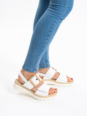 Legs in blue skinny jeans wearing white open-toe sandals with thick soles and large square gold buckles on a white background.
