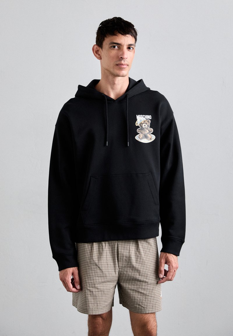 Schwarzer Hoodie aus Baumwolle, mit einer vorderen Tasche und einem Grafikdesign eines Bären mit Hut auf der Brust. Trägt dazu karierte Shorts.