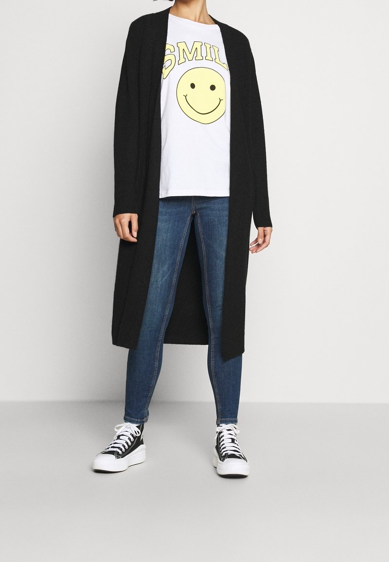 Personne portant un t-shirt blanc avec un visage souriant jaune et le texte « SMILE », un cardigan long noir, un jean skinny bleu et des baskets noir et blanc.