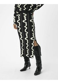 Jupe en tricot noir et blanc avec un motif ondulé, fente latérale, associée à des bottes noires jusqu'aux genoux à bout pointu et talon aiguille.