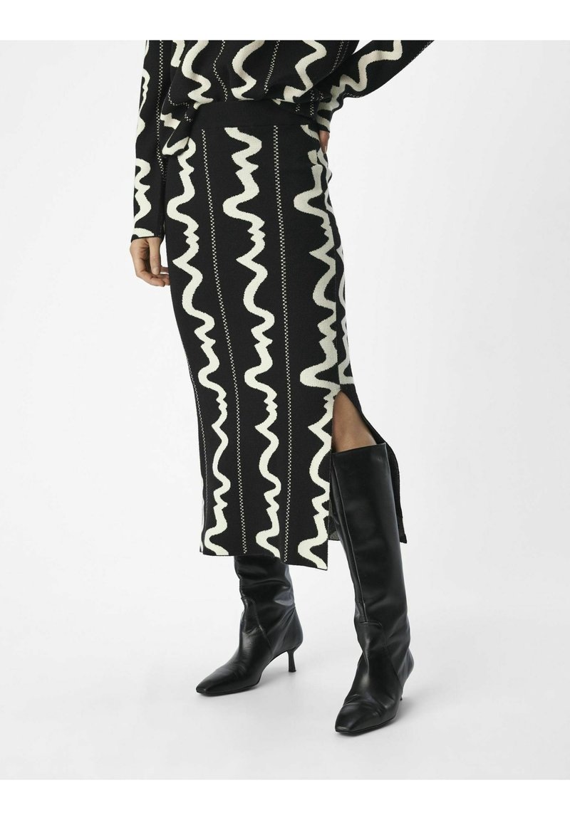 Jupe en tricot noir et blanc avec un motif ondulé, fente latérale, associée à des bottes noires jusqu'aux genoux à bout pointu et talon aiguille.