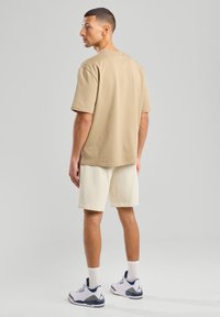 T-shirt a maniche corte color tan, in cotone, dalla vestibilità rilassata; abbinato a pantaloni corti color crema chiaro. Sneakers bianche con dettagli blu. Sfondo neutro.