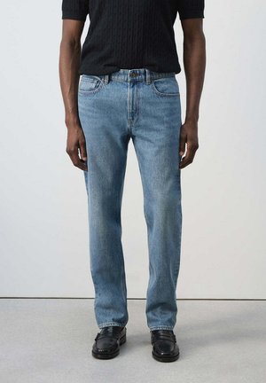 Mann trägt hellblaue gerade Jeans, schwarzes strukturiertes kurzärmeliges Hemd und schwarze Lederloafer und steht vor einem einfarbigen Hintergrund.