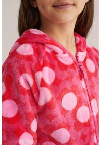 Raudonas ir rožinis taškuotas flisinis džemperis su priekiniais užtrauktukais ir gobtuvu. Minkšta tekstūra, apvalūs raštai skirtingais rožinės atspalviais.