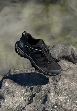 Czarny but trekkingowy The North Face z podeszwą Vibram zawieszony nad skalistym terenem.