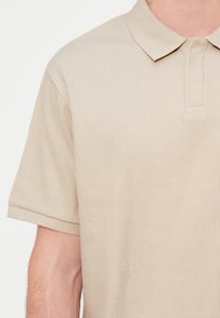 Μπεζ ριγωτό πόλο πουκάμισο με γιακά, διαθέτει κοντό μανίκι με ρεβέρ και κουμπωτό άνοιγμα. Ομαλή υφή, χωρίς ορατά μοτίβα ή διακοσμήσεις.