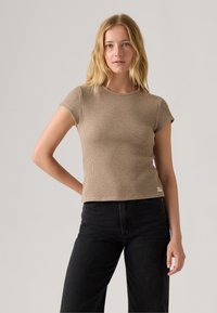 Camiseta marrón de manga corta acanalada con corte recto, cuello redondo y una pequeña etiqueta de logo en el dobladillo. Lleva puesta unos vaqueros negros de cintura alta.