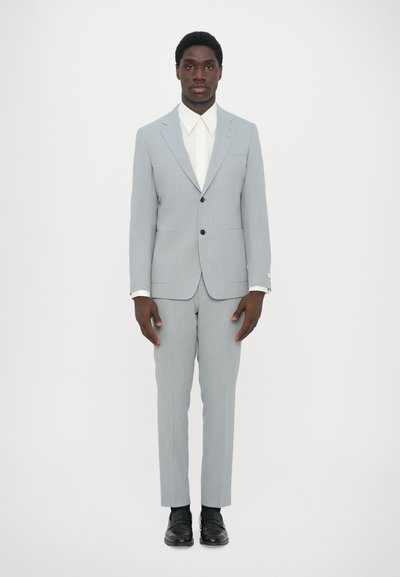 Lys grå jakkesæt med en tætsiddende jakke og lige bukser, parret med en hvid skjorte med knapper. Sorte sko fuldender outfittet.