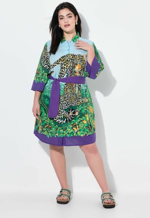 Vestido camisero impreso con un diseño de leopardo en azul, verde y morado, con cintura ajustada y patrones de hojas. Se lleva con sandalias verdes.