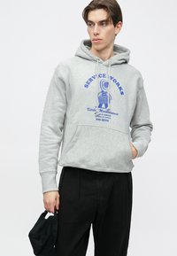 Siva majica s kapuljačom i prednjim džepom, s plavim grafičkim natpisom "SERVICE WORKS." Model nosi crne hlače i drži kapu.