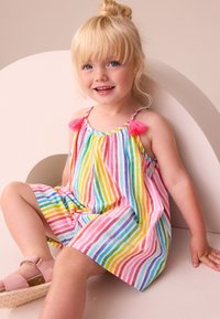 Kleurrijke gestreepte jurk met dunne bandjes, roze kwastjes en een losse pasvorm. Het materiaal lijkt lichtgewicht en textuurrijk. Roze sandalen met een sleehak.