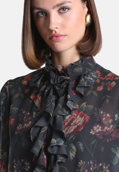 Jeune femme aux cheveux courts et bruns portant des boucles d'oreilles en or et un chemisier sombre à motifs floraux avec un col à volants et des détails à l'avant.