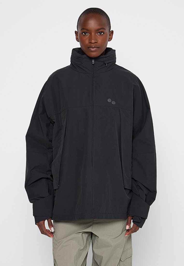 Schwarze, oversize Jacke mit hohem Kragen, mittigem Reißverschluss und dezenten Logo-Akzenten. Aus leichtem Material mit matter Textur gefertigt.