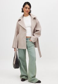Abrigo de lana beige claro con solapas recortadas, mangas oversize y lazos laterales. Lleva puesto sobre una blusa blanca y vaqueros de pierna ancha verdes. Bolso negro.