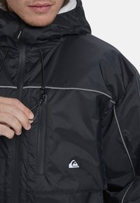 Black waterproof jacket with a hood, gray accents, zippered pocket, and drawstring detail. Features a logo on the left chest area.
