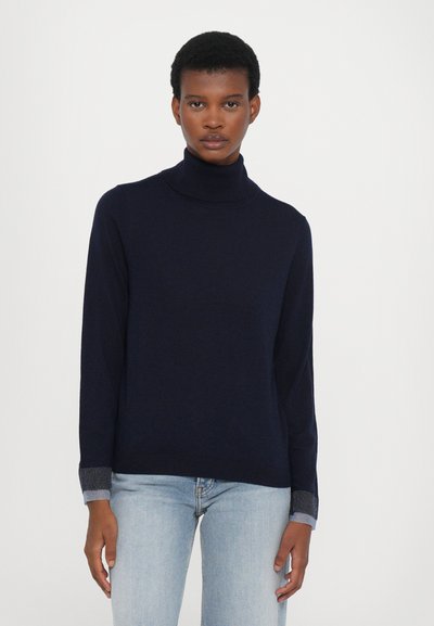 Merinovillane turtleneck-neule, joka on valmistettu pehmeästä materiaalista, pitkät hihat ja harmaat ribatut hihansuut, yhdistettynä vaaleansinisiin farkkuihin.