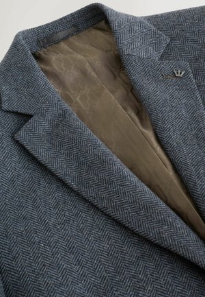 Grå sildebensmønstret blazer med skrå kraver, med en glat brun foring og en lille metalemblem på reversen. Tekstureret stoffinish.