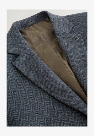 Grå sildebensmønstret blazer med skrå kraver, med en glat brun foring og en lille metalemblem på reversen. Tekstureret stoffinish.