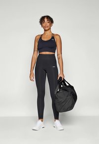 Crni sportski crop top i tajice visokog struka od rastezljivog materijala, uparen s crnom torbom za teretanu i bijelim tenisicama.