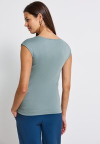 Top ajustado verde claro con mangas cortas y escote redondo. Textura suave, visto desde la parte de atrás, combinado con pantalones azules.