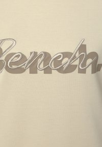 Crème-kleurige stof met het woord "Bench" in grijze en zilveren borduurwerk, met verschillende lettertypen en texturen.