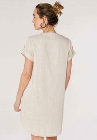 Robe chemise en coton beige avec des manches courtes roulées et un ourlet arrondi, présentant un motif tissé texturé. Design simple et dépouillé.