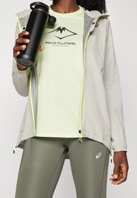 Persona vestida con camiseta "ASICS FUJITRAIL" verde claro, leggings gris-verde, chaqueta gris claro con detalles en cremallera neón, sosteniendo una botella de agua negra.